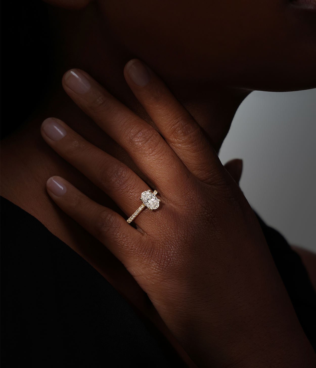 A model wearing an 18ct yellow gold diamond engagement ring, with an oval cut laboratory grown diamond at the centre and diamond-set shoulders.