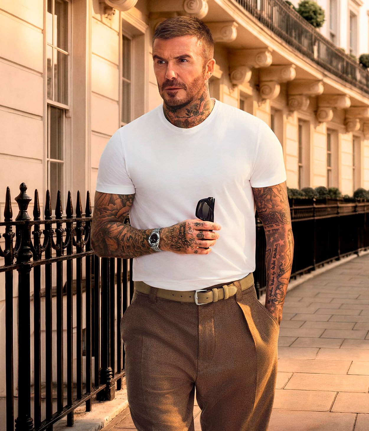 David Beckham wearing a white t-shirt with tattoos on his hands and wrist, holding a pair of sunglasses