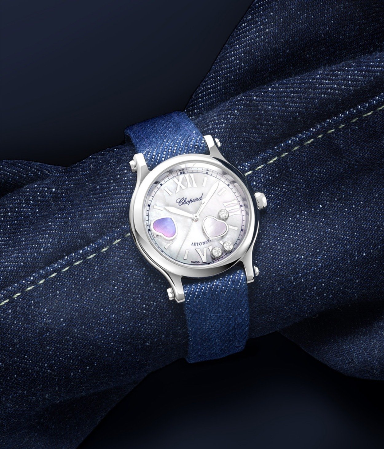 Close-up of a person's clasped hands near their chin, wearing a Chopard wristwatch with a black leather strap and Roman numeral dial, alongside a gold bracelet; soft-focus light background.