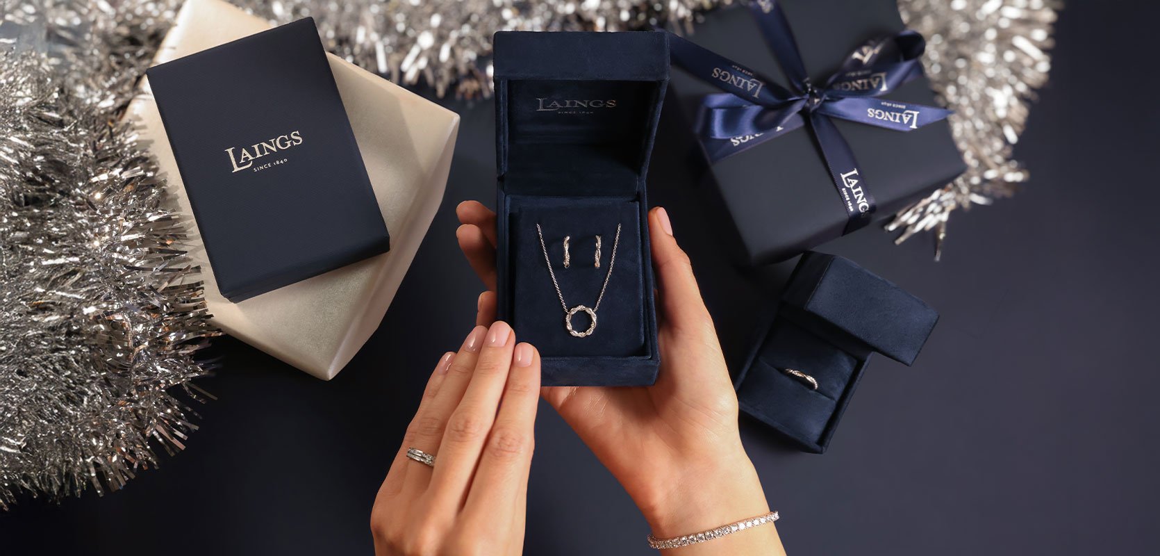 A ladies hand holding a gift box, with a jewellery set including an 18ct yellow gold necklace and earrings.