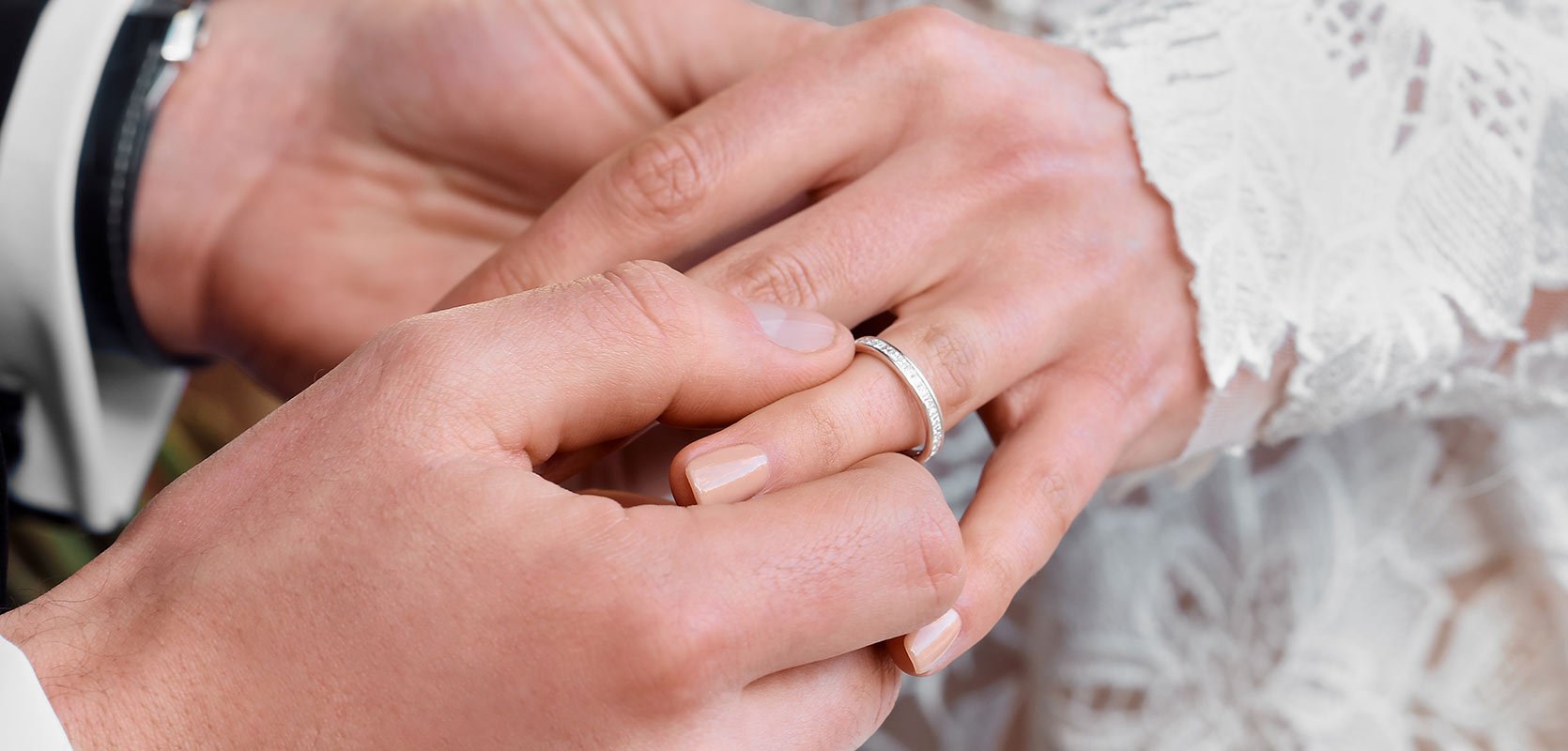 A groom places a diamond-set wedding band onto the brides ring finger.