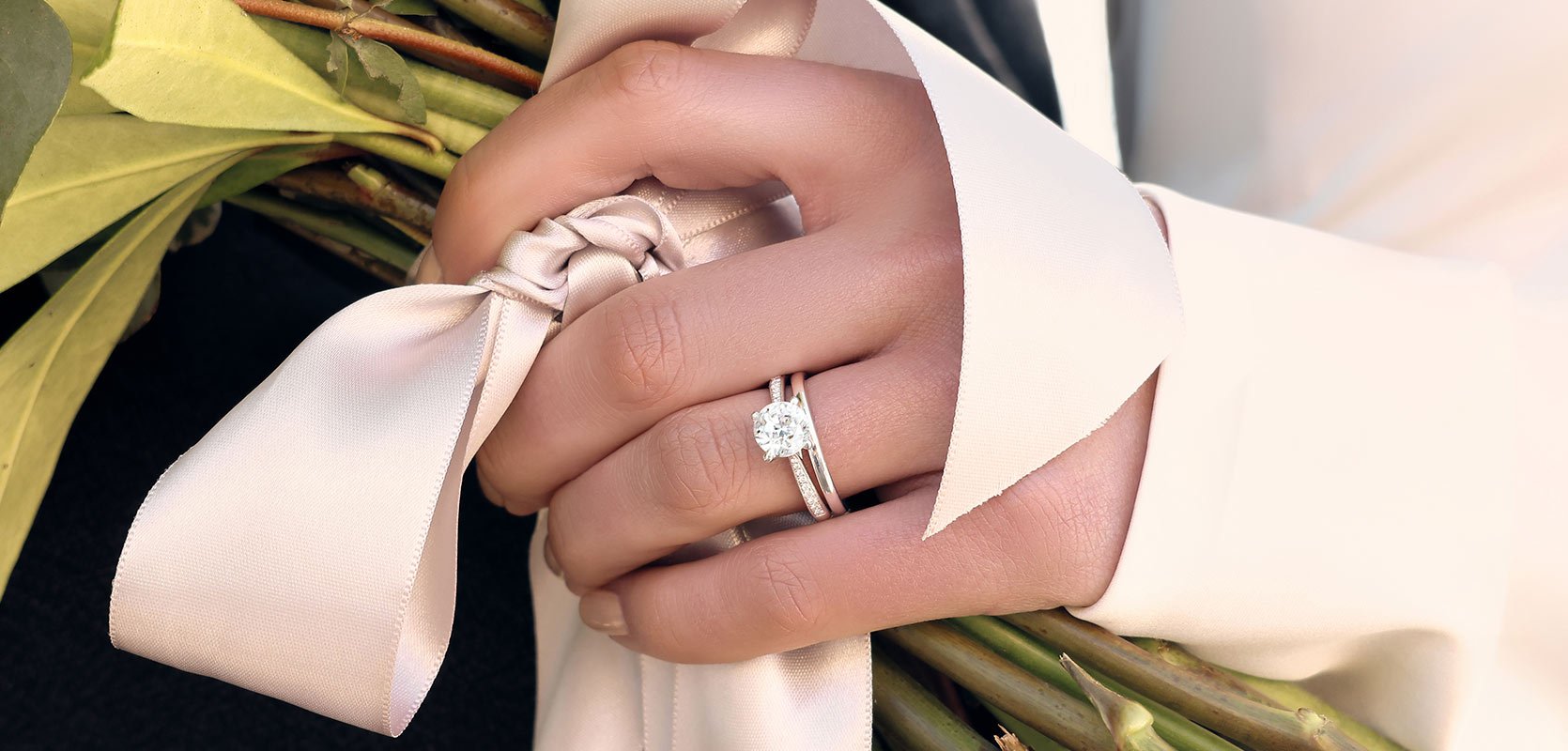A bride clutches a bouquet of flowers, whilst displaying a solitaire diamond engagement ring with diamond shoulders, and a plain platinum band.