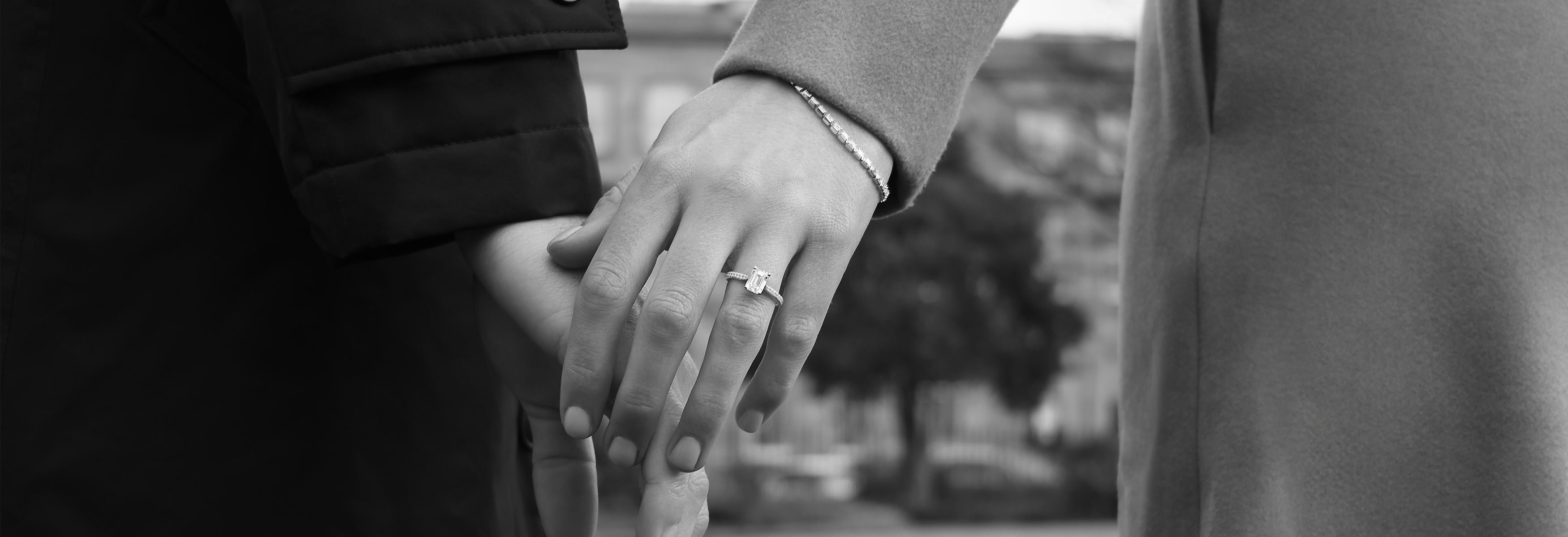 A couple holding hands in black and white, with a focus on the emerald cut engagement ring and diamond line bracelet.
