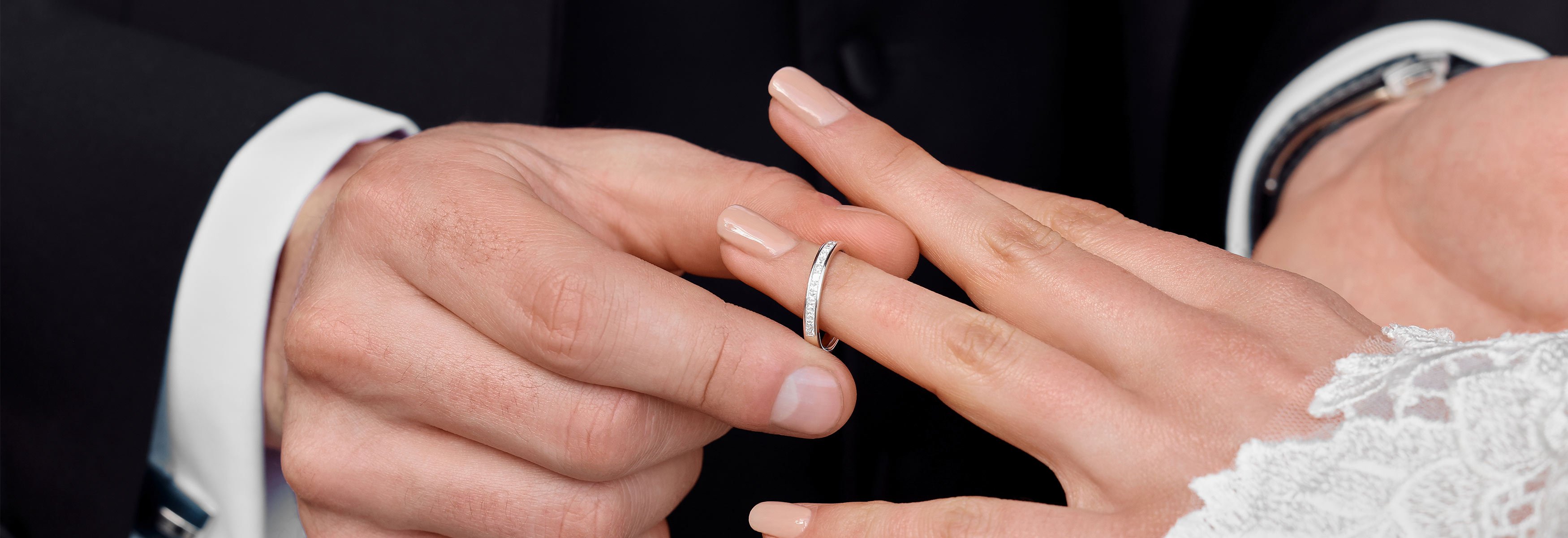 A groom places a diamond-set wedding band onto the brides ring finger.