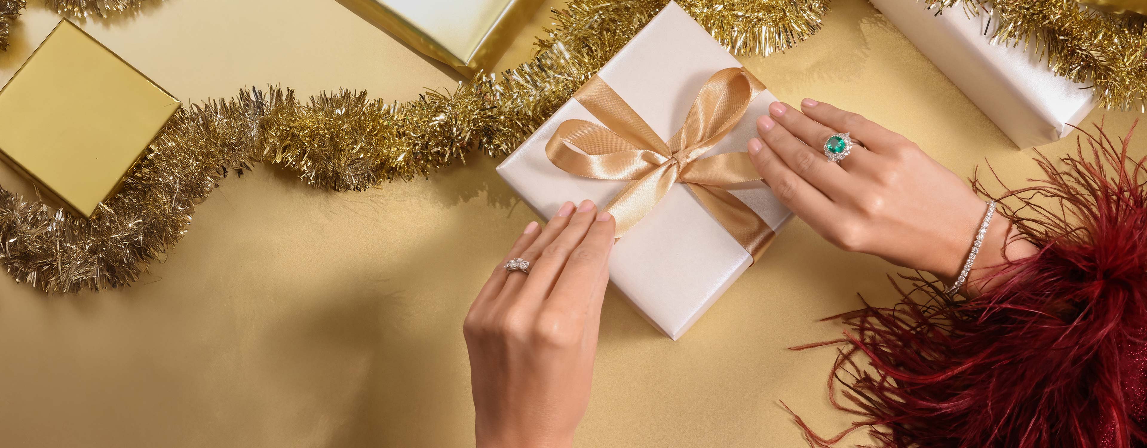 A ladies hand holding a gold present box, with a gold ribbon. Wearing two rings, one emerald and one full diamond.