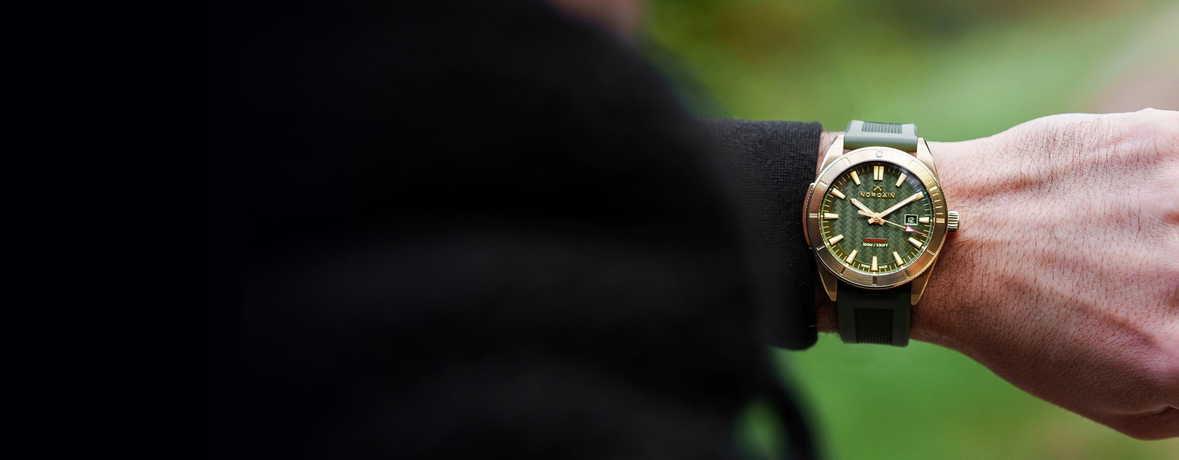 A mans wrist wearing the new NORQAIN Wild One timepiece in olive green with yellow gold accents and a rubber strap.