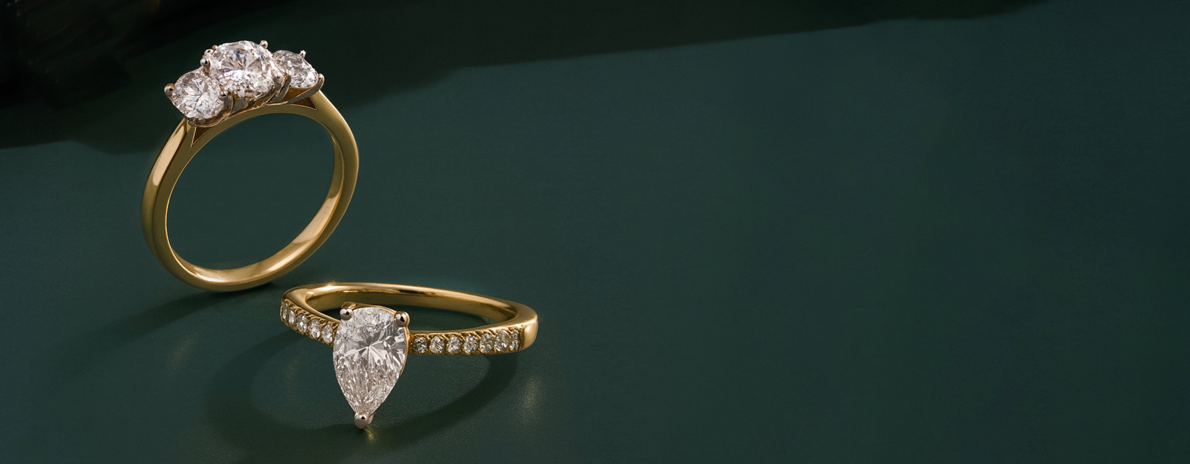Two engagement rings in 18ct yellow gold, one with three oval cut diamonds and the other with a centre pear cut diamond and diamond-set shoulders.
