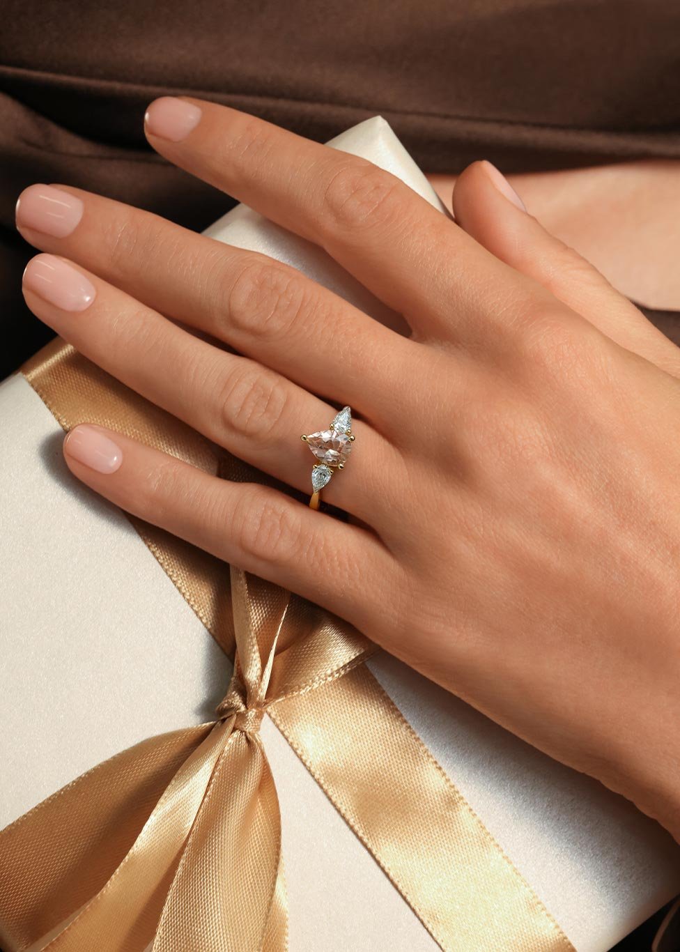 A ladies hands wearing a morganite and diamond three stone ring, whilst wrapping a silver present with a gold bow.