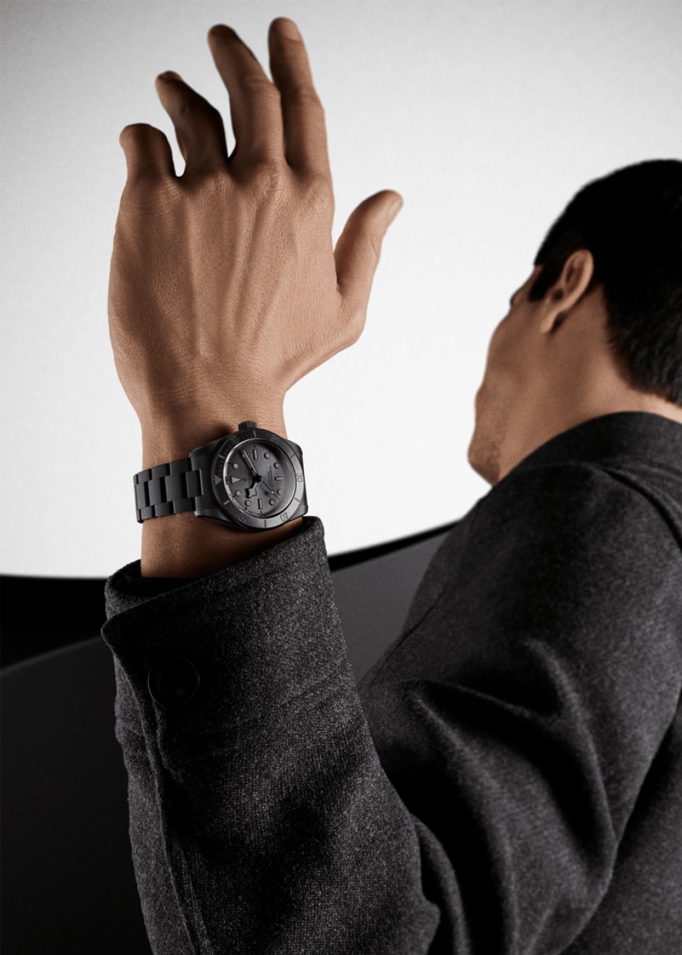 A person seen from behind raises a hand wearing a black metal wristwatch, with the watch face clearly visible against a minimalist light background.