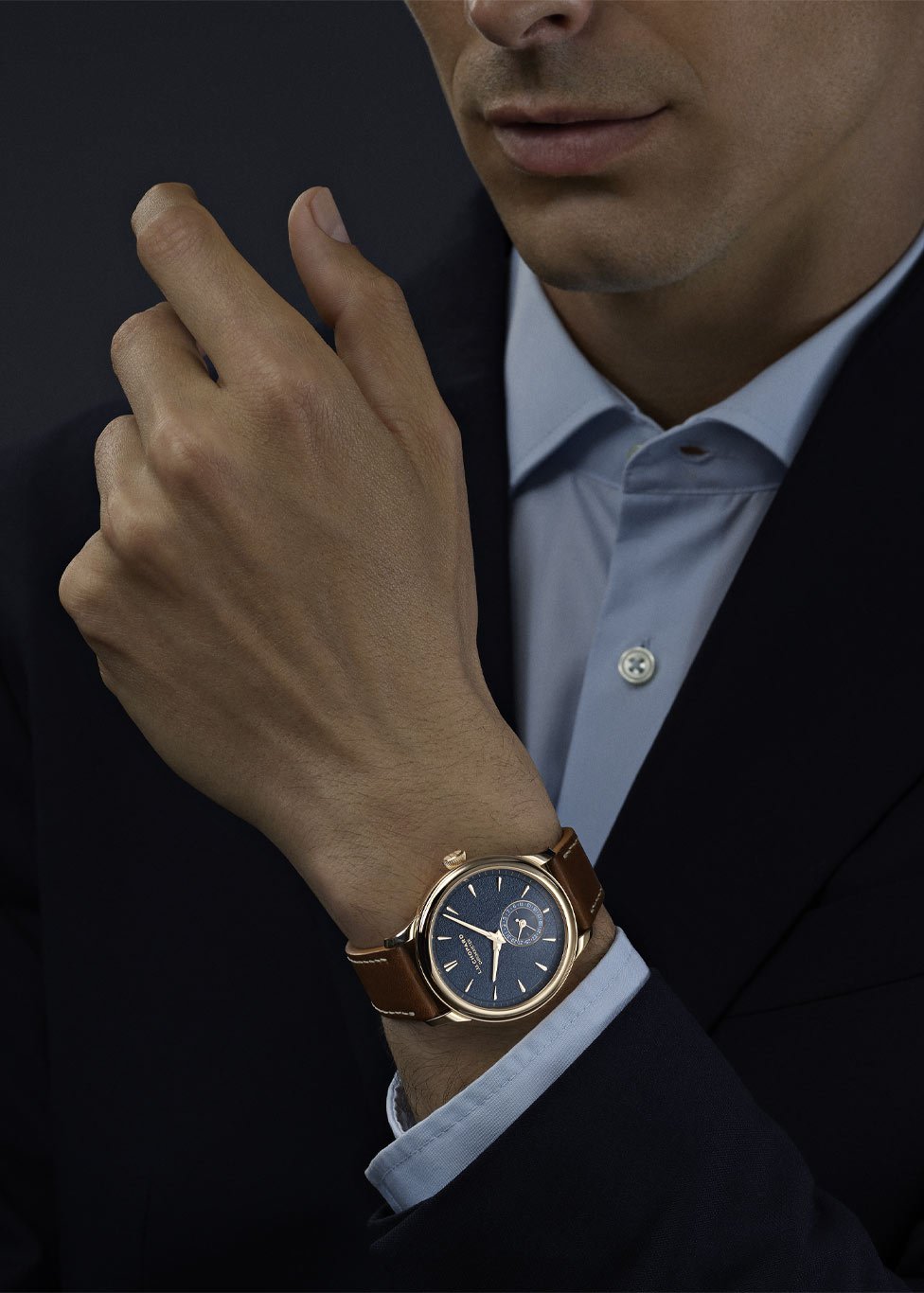 Luxury wristwatch dispalyed on a wrist with a blue dial and small seconds sub-dial, in a gold case with a brown leather strap. The gent is dressed in a dark suit jacket with a light blue shirt. The background is dark and minimalistic.