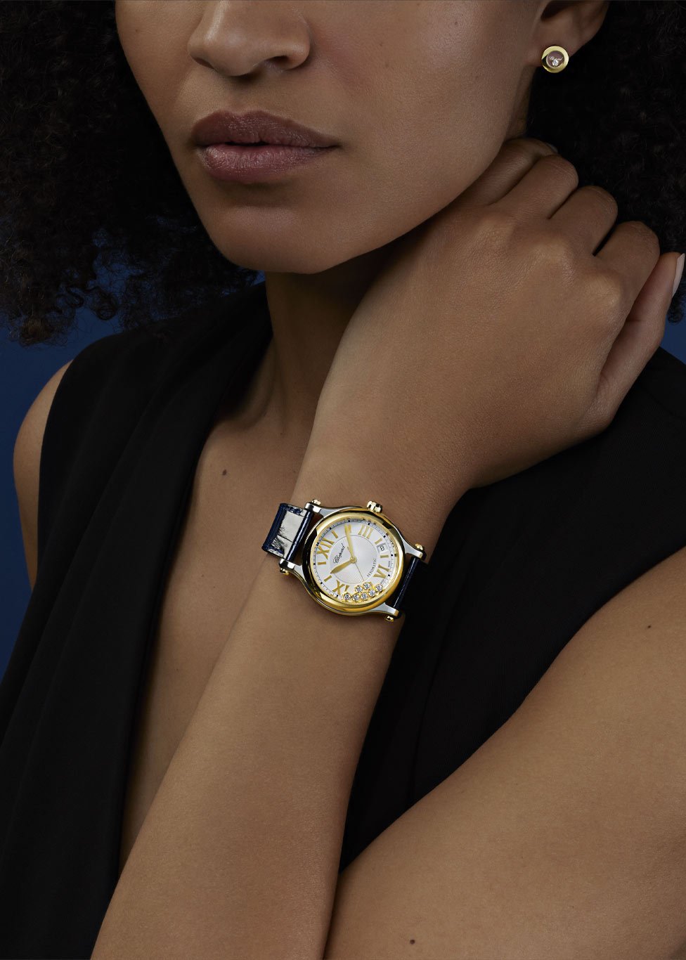 A close-up of a woman's wrist showcasing a watch with a round gold case and a white dial featuring elegant hour markers and a glossy dark blue leather strap. The background is a deep blue, creating a sophisticated and elegant setting.