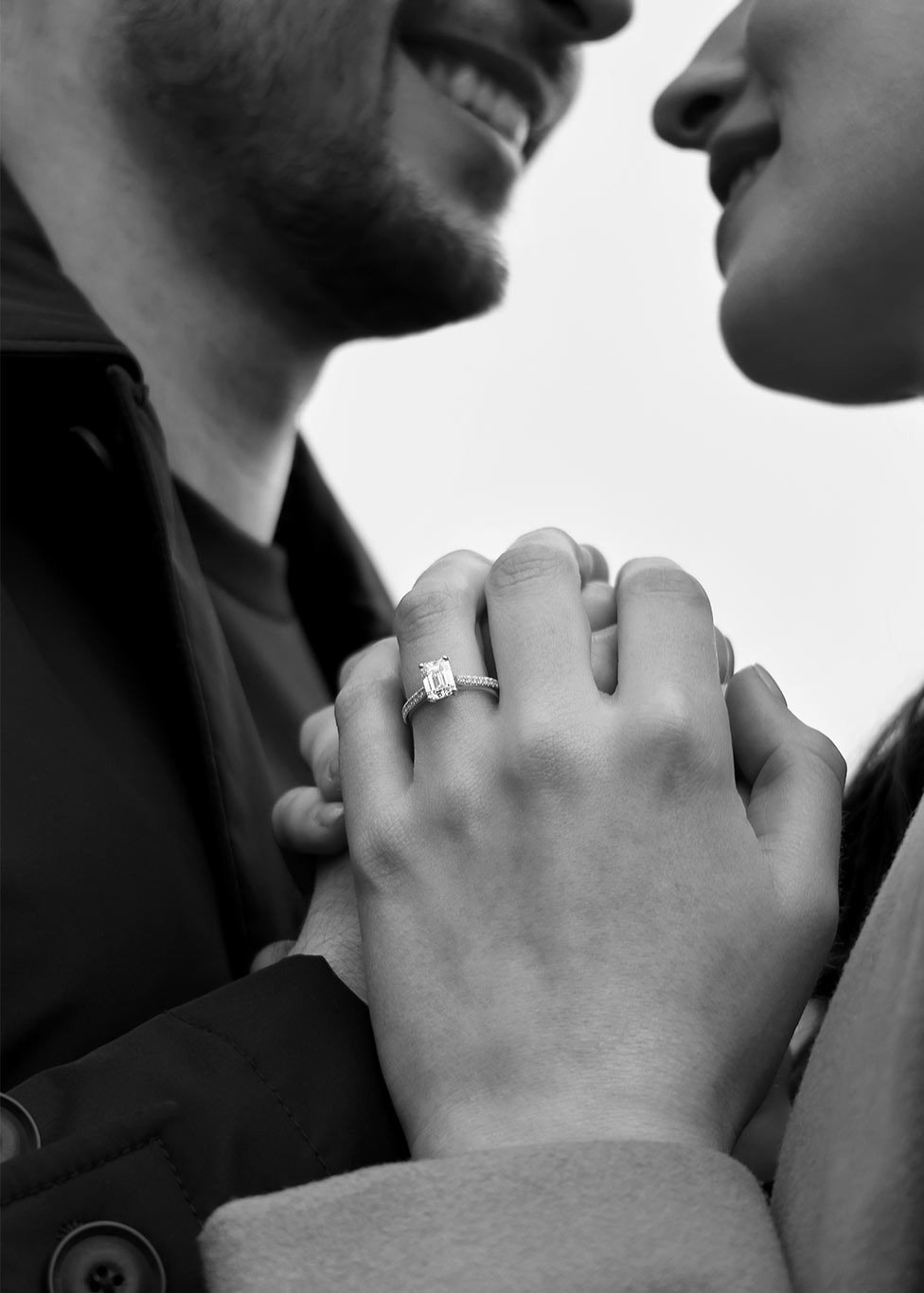 A black and white image of a couple, with focus on her diamond engagement ring that features an emerald cut diamond at the centre, with diamond set shoulders.