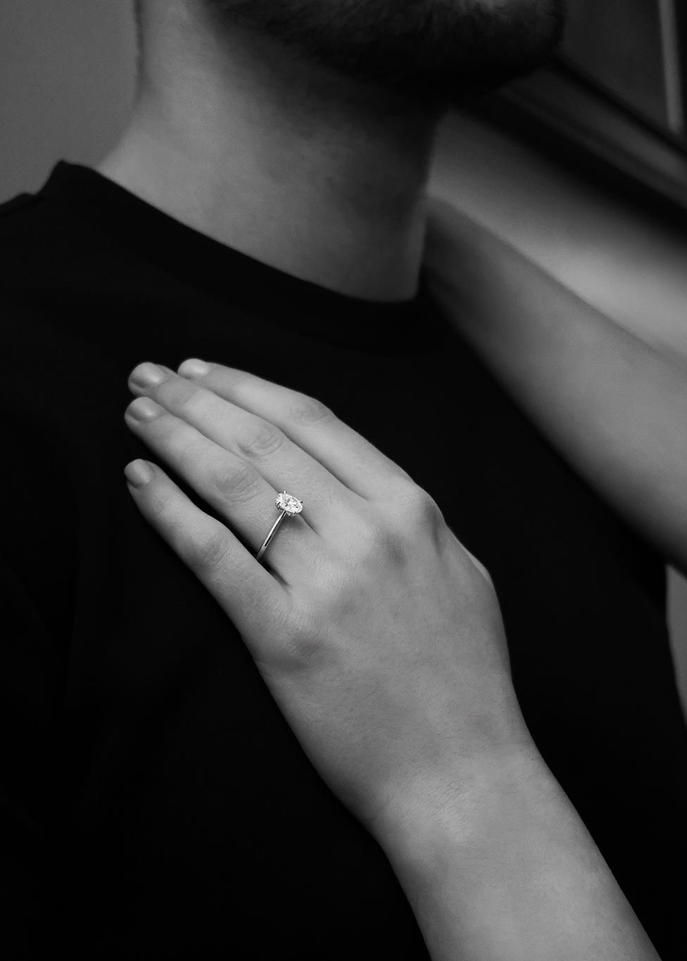 A black and white image of a couple, with focus on her diamond engagement ring that features an oval cut diamond at the centre.