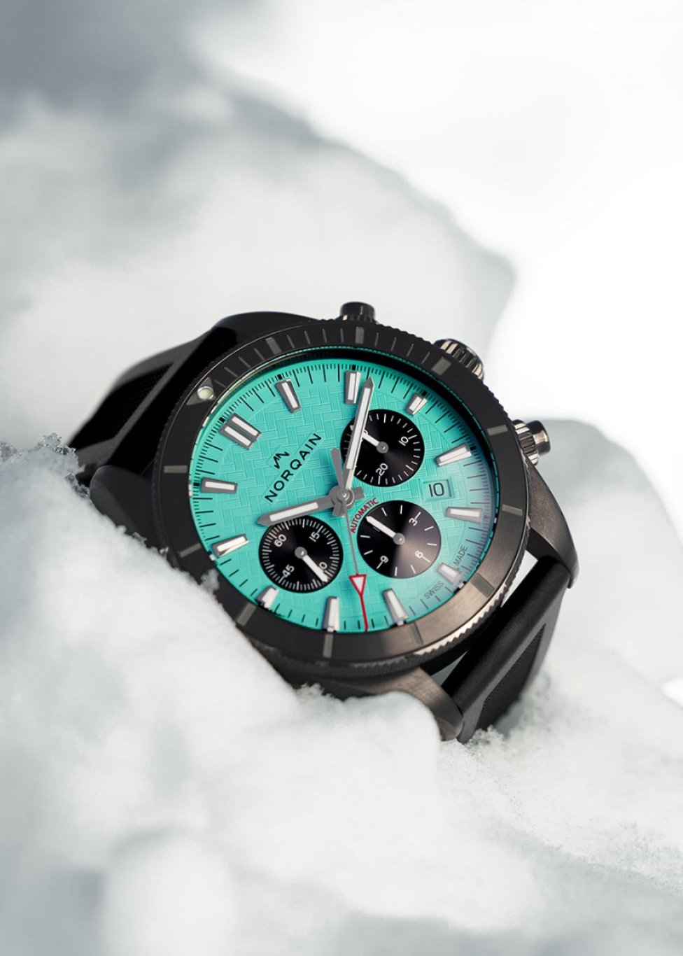A NORQAIN watch with a turquoise blue chronograph dial, with a black bezel, case and rubber strap, sitting within snow. 