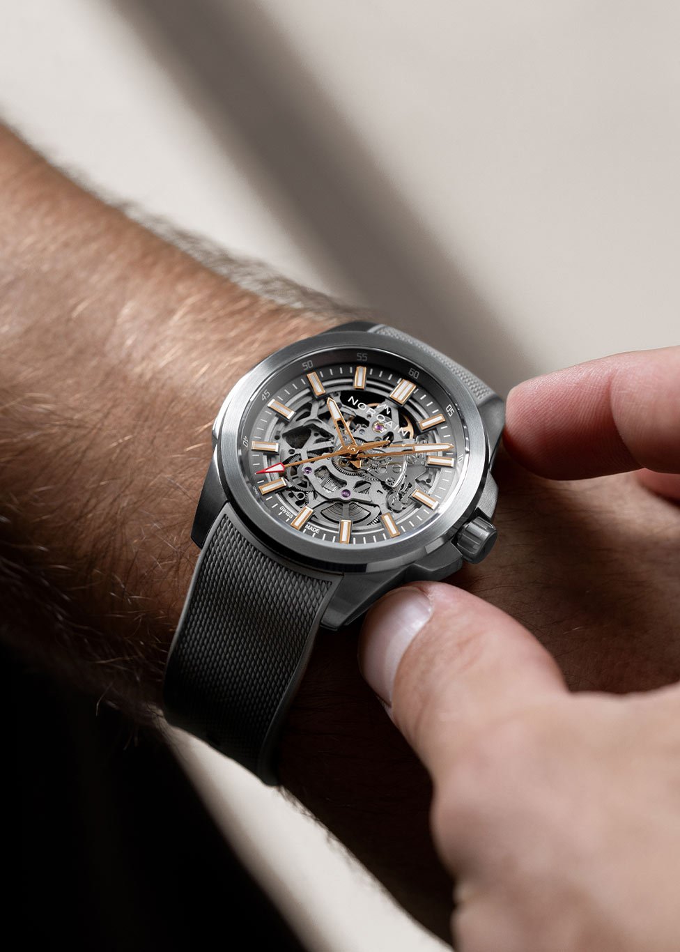 A close-up of a luxury wristwatch with a skeleton dial the mechanical movement. The watch features a round stainless steel case, bold hour markers, and a textured grey strap.  The watch is worn on the wrist, set against a softly lit neutral background.