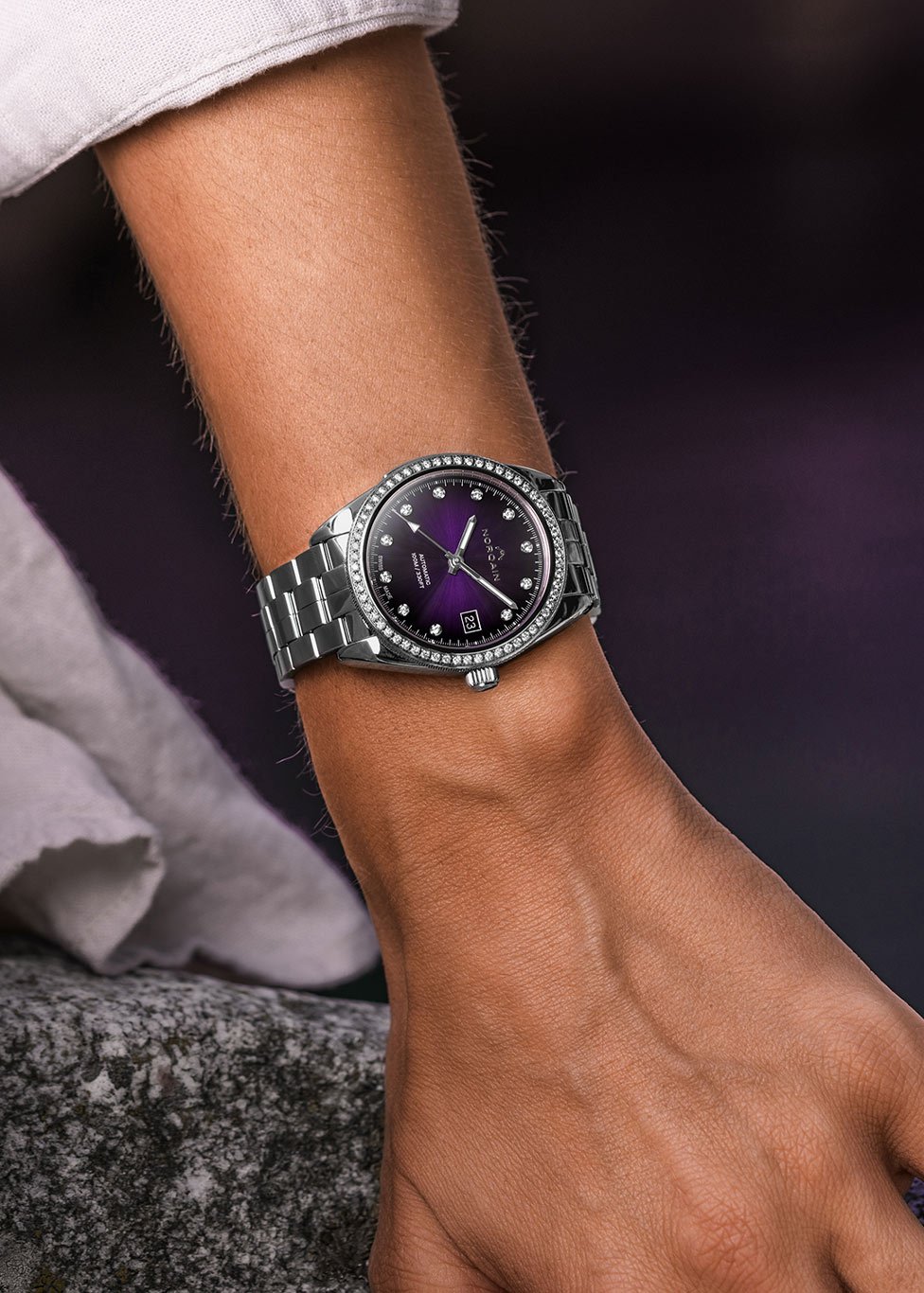 Close-up of a wrist wearing a luxury stainless steel watch with a deep purple dial, diamond hour markers and a bezel. The watch features a date display at 6 o&rsquo;clock and a metal bracelet. The background is softly blurred in dark purple tones.