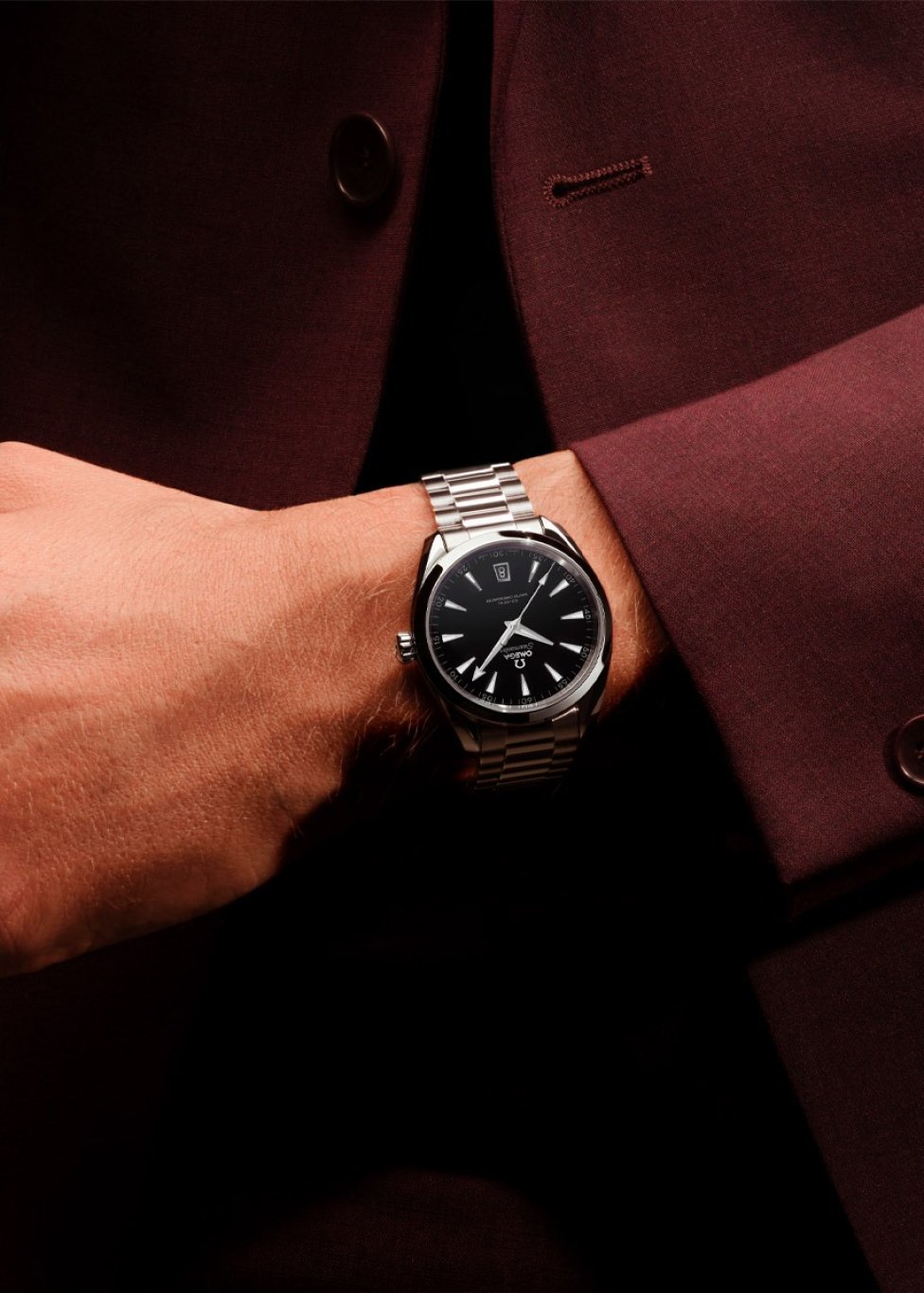 Close-up of a wrist wearing a luxury watch with a black dial and silver metal bracelet. The watch features a date window and sleek hour markers. The person is dressed in a deep burgundy suit jacket.