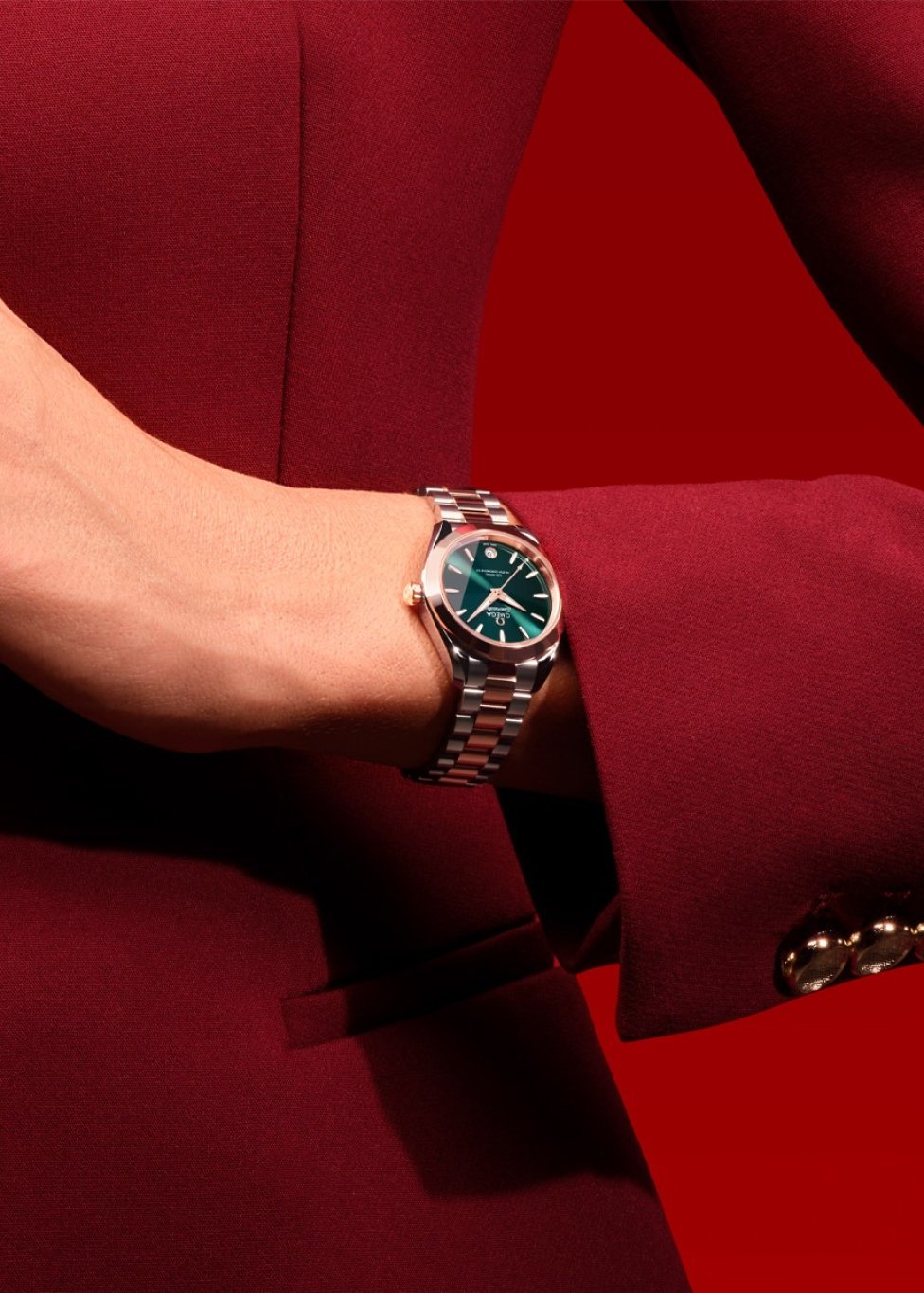 Close-up of a wrist wearing a luxury watch with a green dial and silver metal bracelet featuring rose gold accents. The person is dressed in a deep burgundy suit jacket with gold buttons, set against a bold red background for a festive.